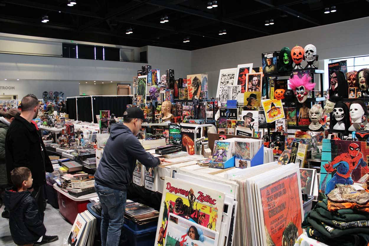 People browse a booth filled with posters, books, and masks at a convention.