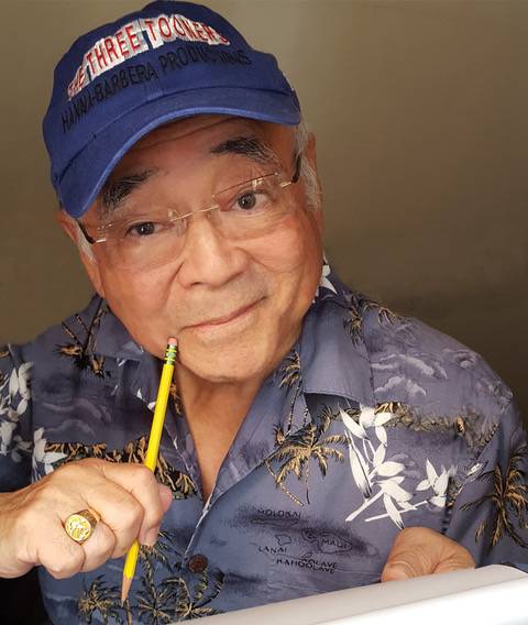 An older man wearing glasses and a blue cap holds a pencil and smiles. He is wearing a patterned shirt and appears to be drawing or writing.