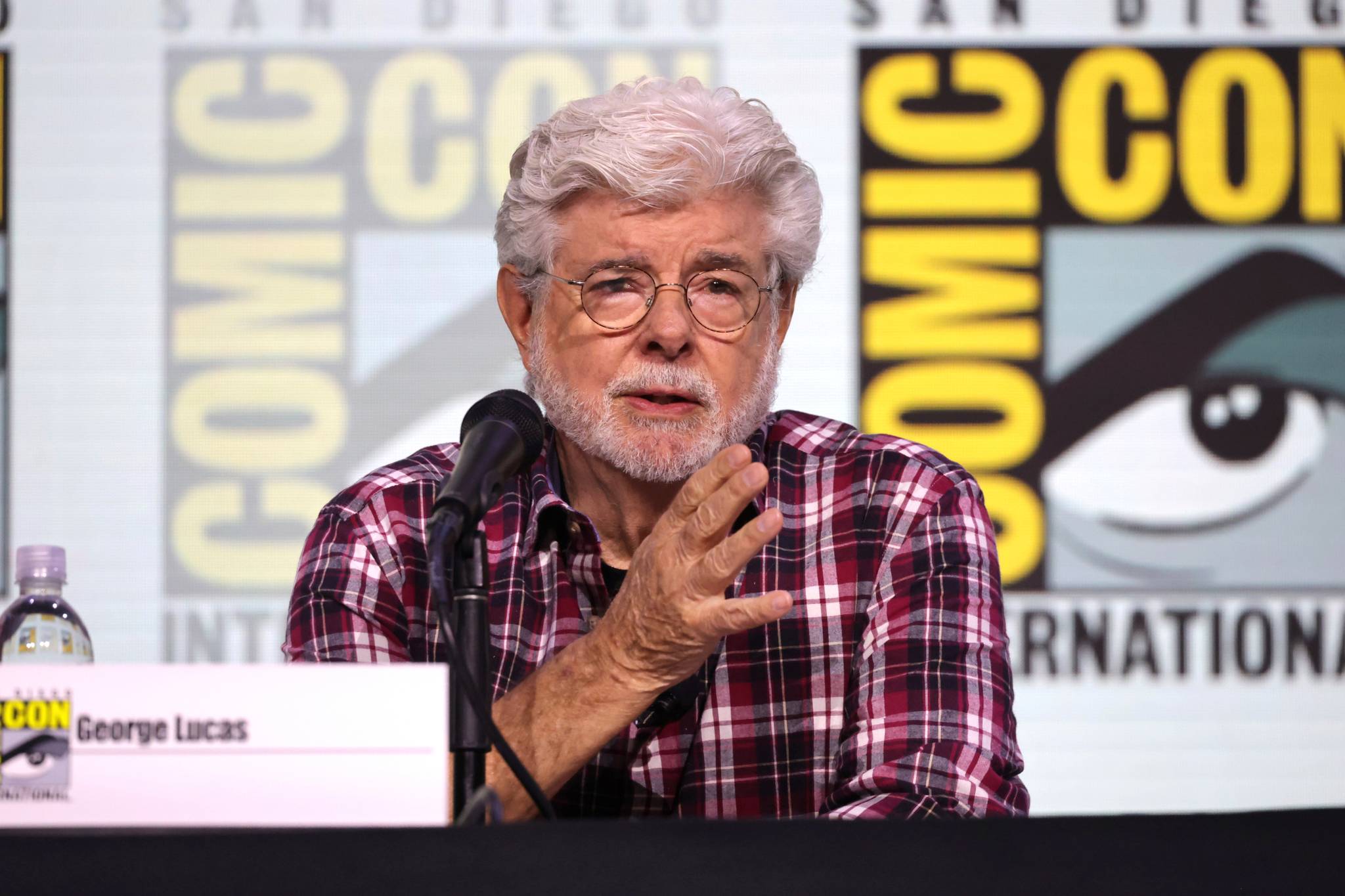 An older man with gray hair and a beard speaks at a Comic-Con panel, seated at a table with a microphone and nameplate reading "George Lucas." Comic-Con logos are in the background.