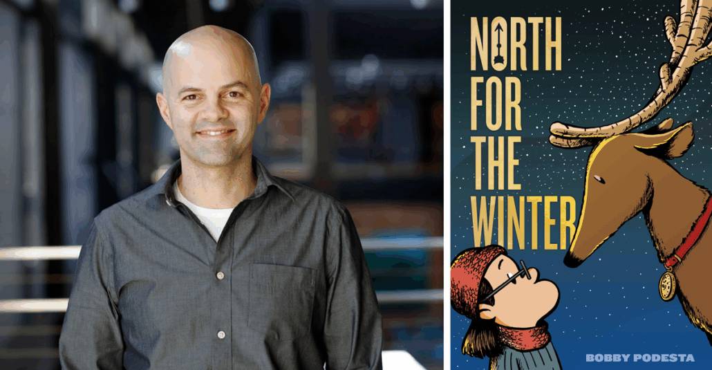 A smiling man stands indoors next to a poster titled "North for the Winter," featuring a reindeer and a child in winter clothing.