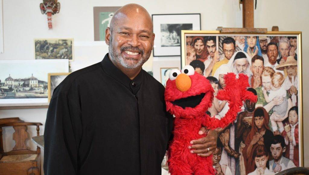 A man smiles while holding the Elmo puppet, standing in front of a Norman Rockwell painting and other framed artwork.