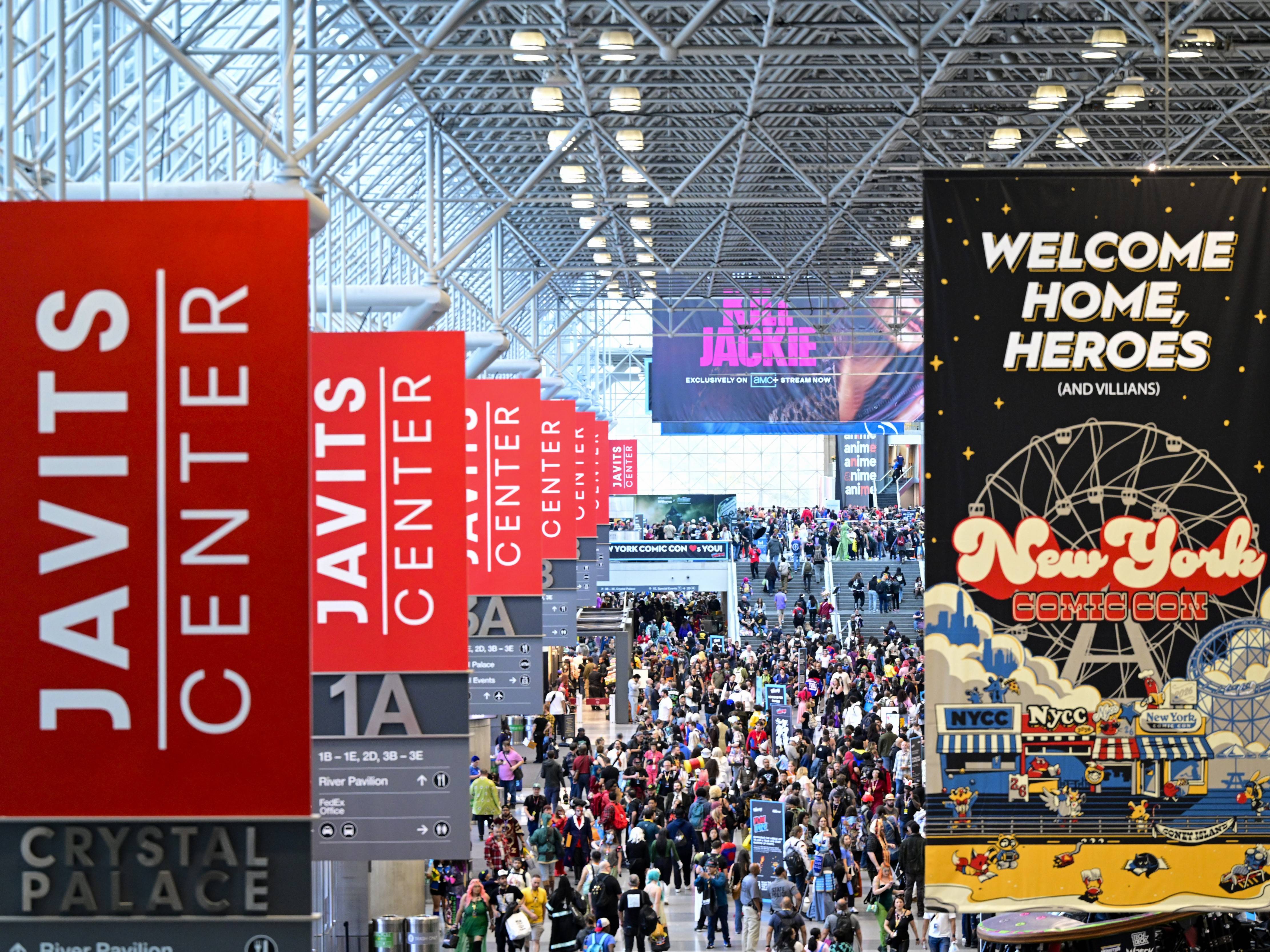 A crowded Javits Center atrium during New York Comic Con, with red vertical banners and a large event sign hanging overhead.